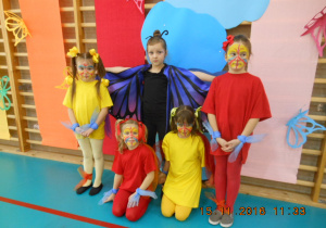 Na tle tęczy stoi pięć dziewczynek w kolorowych strojach, jedna z nich jest przebrana za motyla.